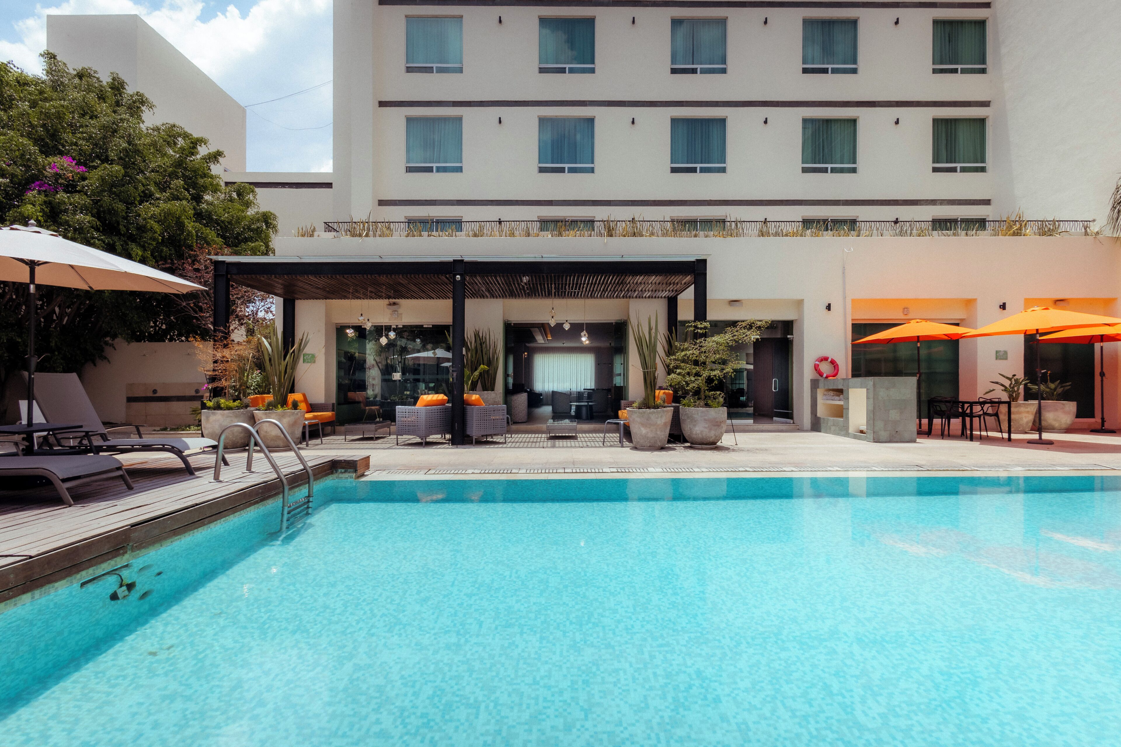 Zona de piscina hotelera con área de estar cubierta, sillas de descanso y sombrillas.
