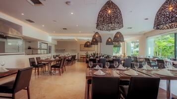 Un comedor amplio con varias mesas dispuestas para los huéspedes, iluminación decorativa en el techo y vistas a zonas verdes al exterior.