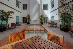 Un patio con una fuente central, rodeado de bancos y sillas de madera, y plantas en macetas.