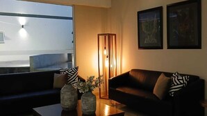 Una sala de estar moderna con un sofá, una mesa de centro y sillones. Se observan obras de arte enmarcadas en la pared y un ventanal con vistas al exterior.