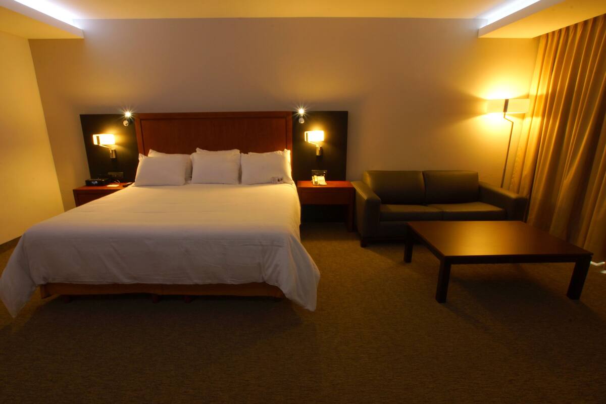 Una habitación de hotel con una cama grande, un sofá, una mesita y dos mesitas de noche con lámparas.