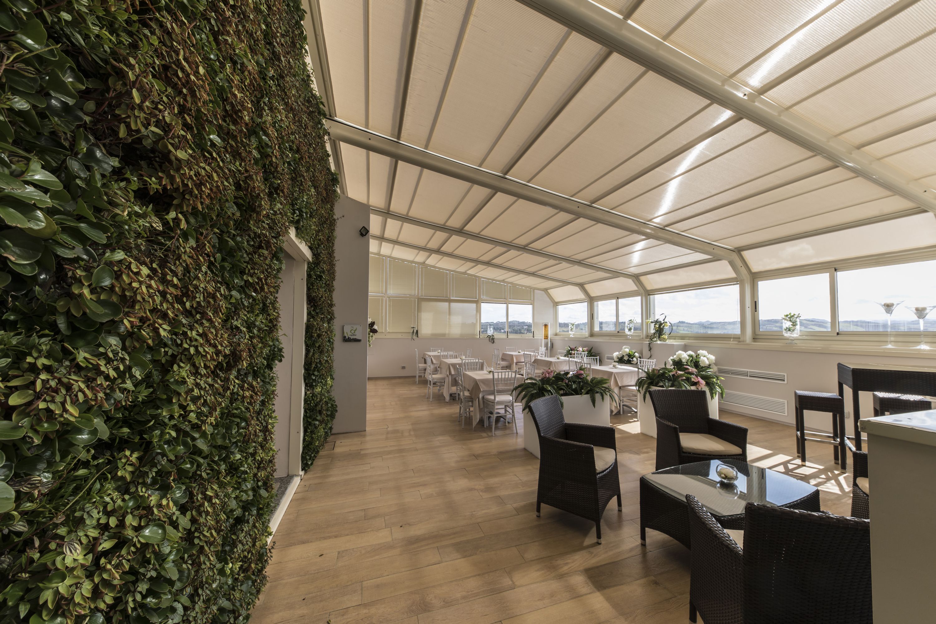 Un espacio interior amplio con techo alto y ventanas grandes, con una pared cubierta de vegetación y varias mesas y sillas dispuestas para comer o conversar.