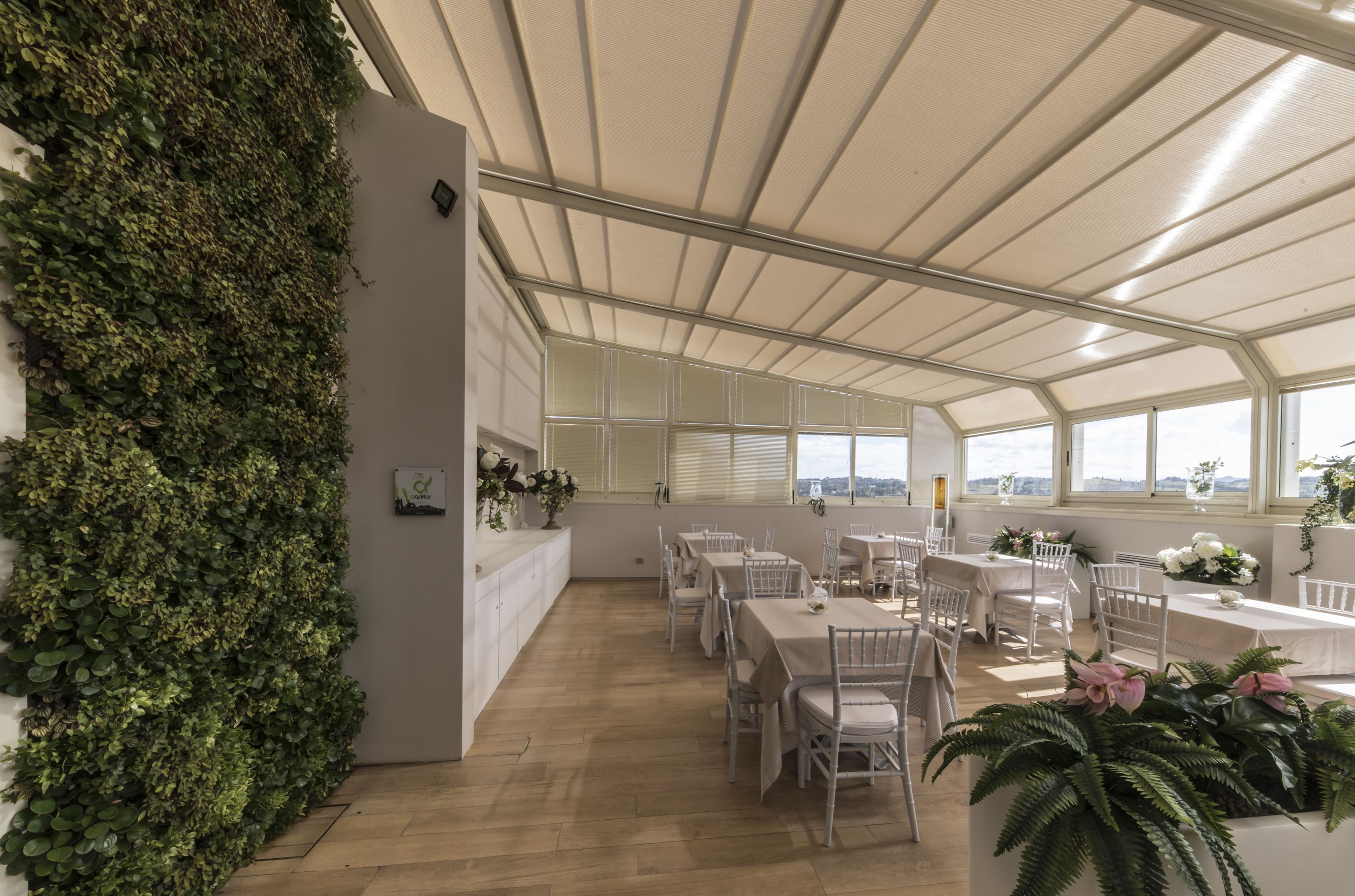 A spacious indoor dining area with white tables and chairs, a high ceiling with a translucent roof, and a large wall covered in greenery.