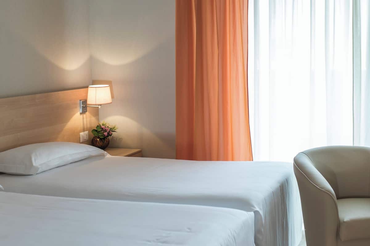 A hotel room with two beds, a nightstand with a lamp, a chair, and a window with curtains.