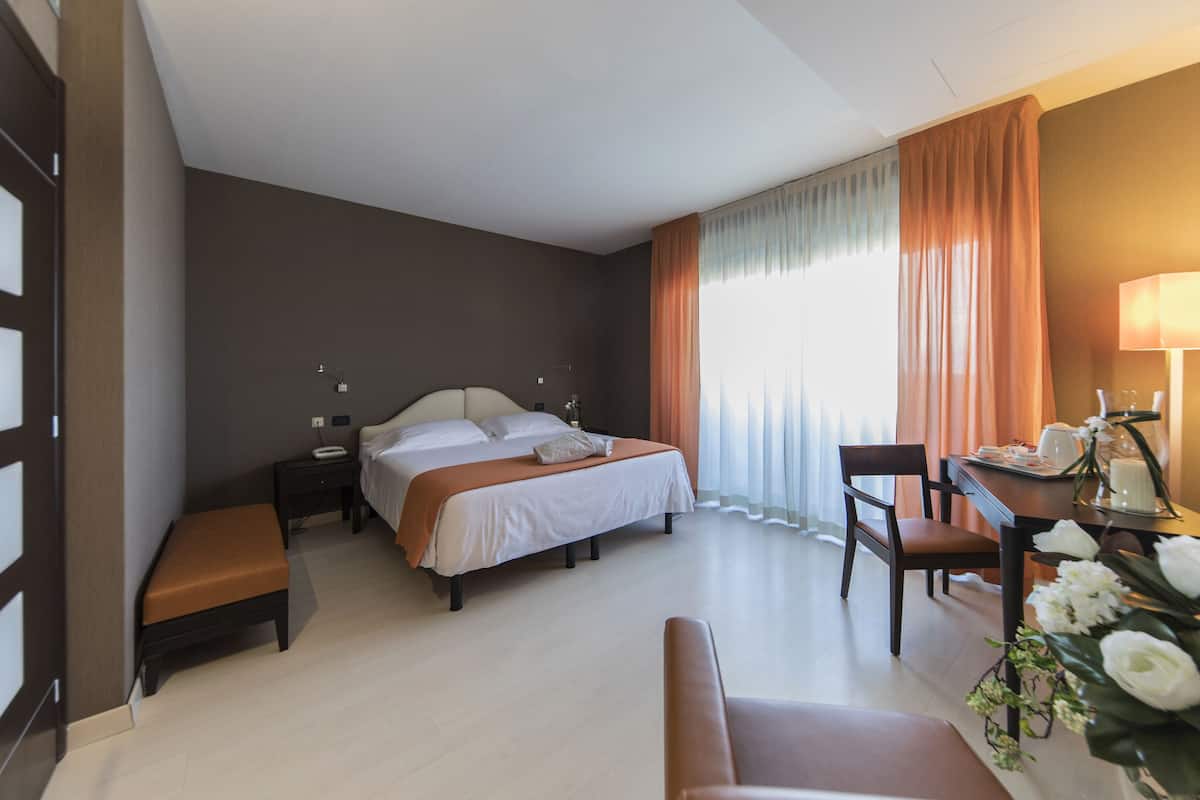 A hotel room with a bed, bedside tables, a desk with a lamp, a chair, and a bench.