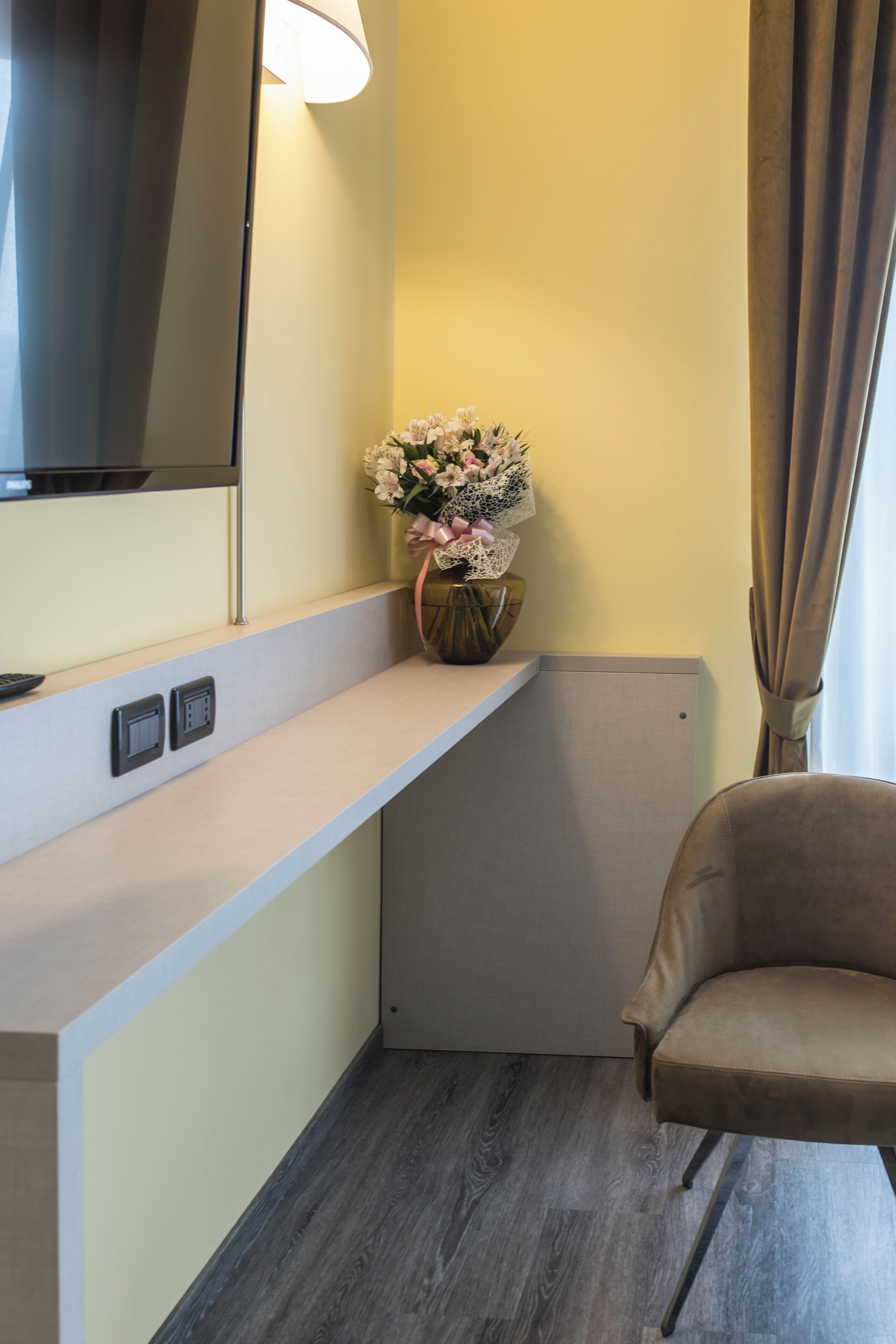 A hotel room with a light-colored wall, a dark wood floor, a beige chair, a small table with a vase of flowers, a flat-screen TV mounted on the wall, and a power outlet on the wall.