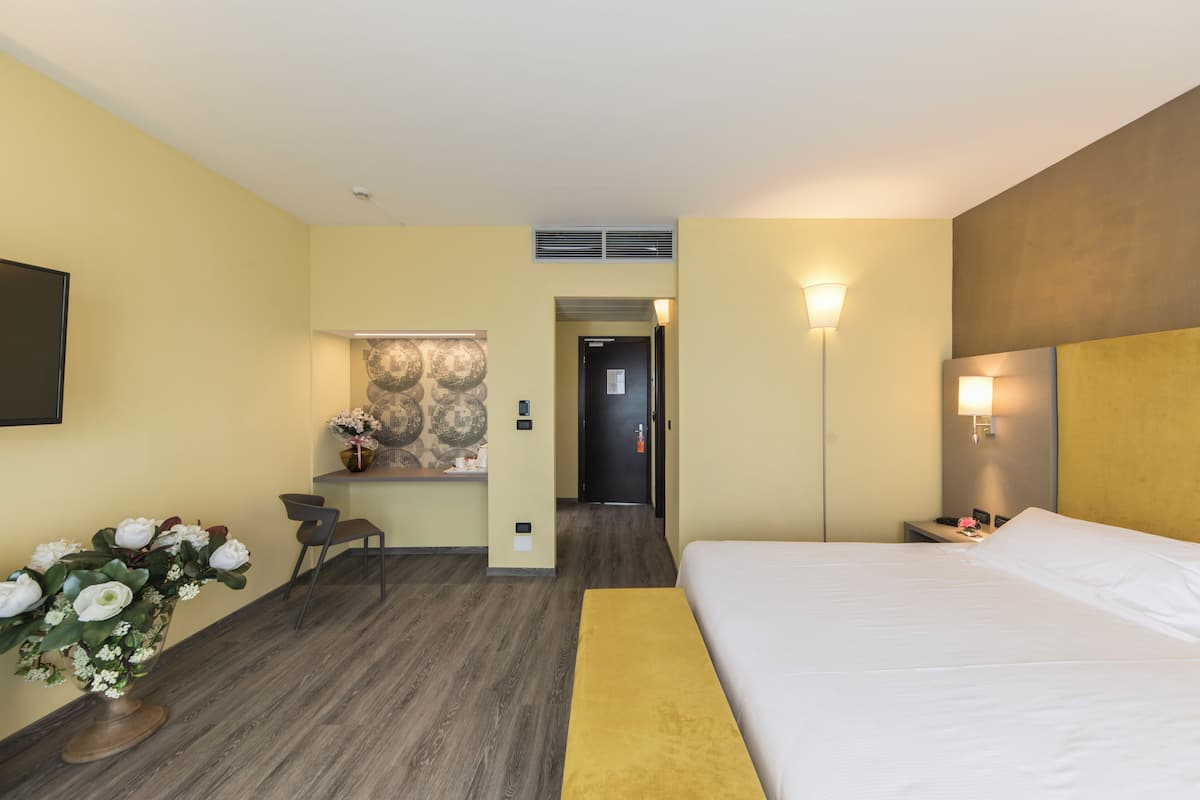 A hotel room with a bed, a desk, a chair, a vase of flowers, and a wall-mounted TV.
