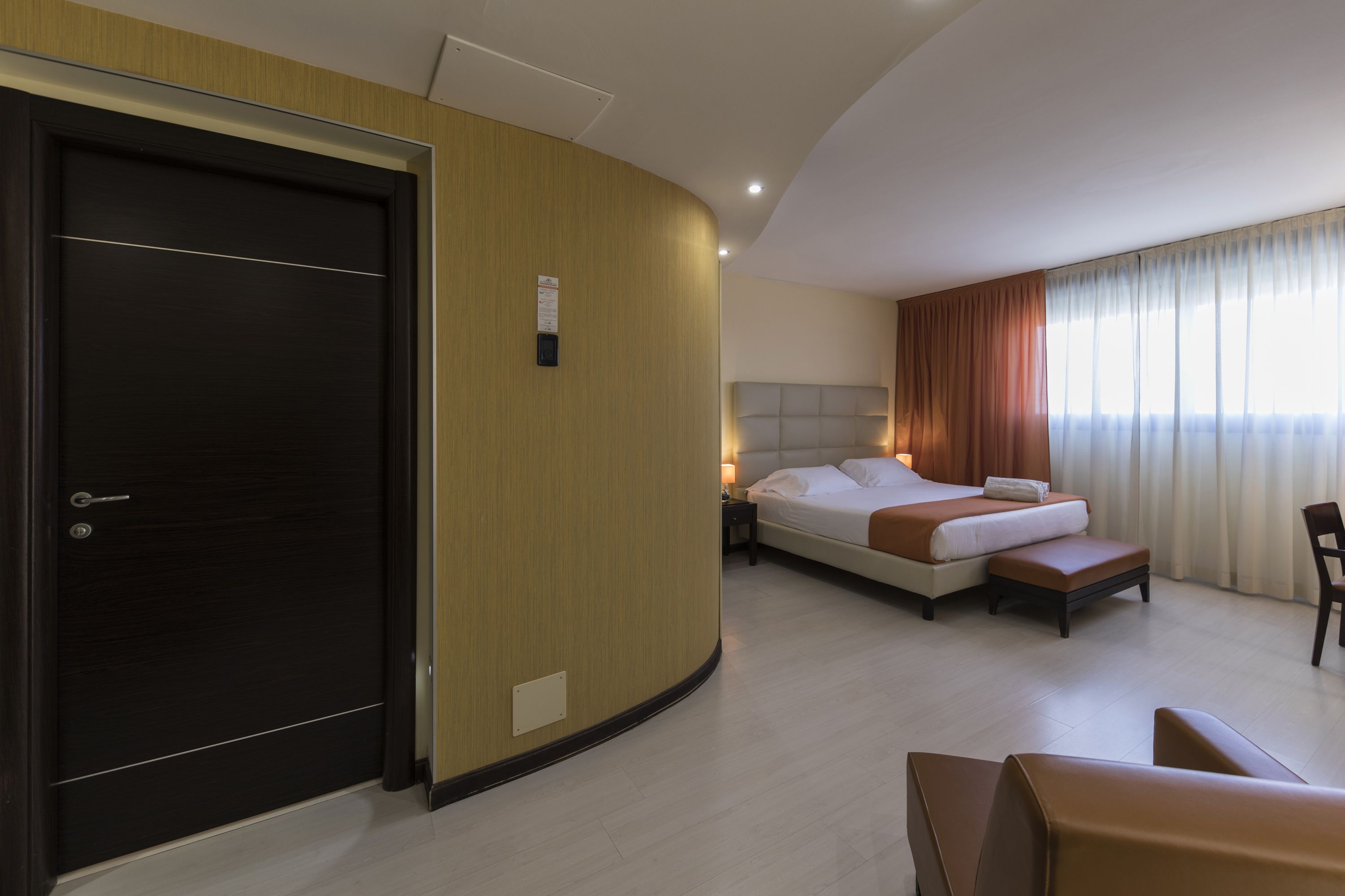 Una habitación de hotel con una puerta de madera oscura, una cama con cabecera blanca, una mesita de noche y una silla.