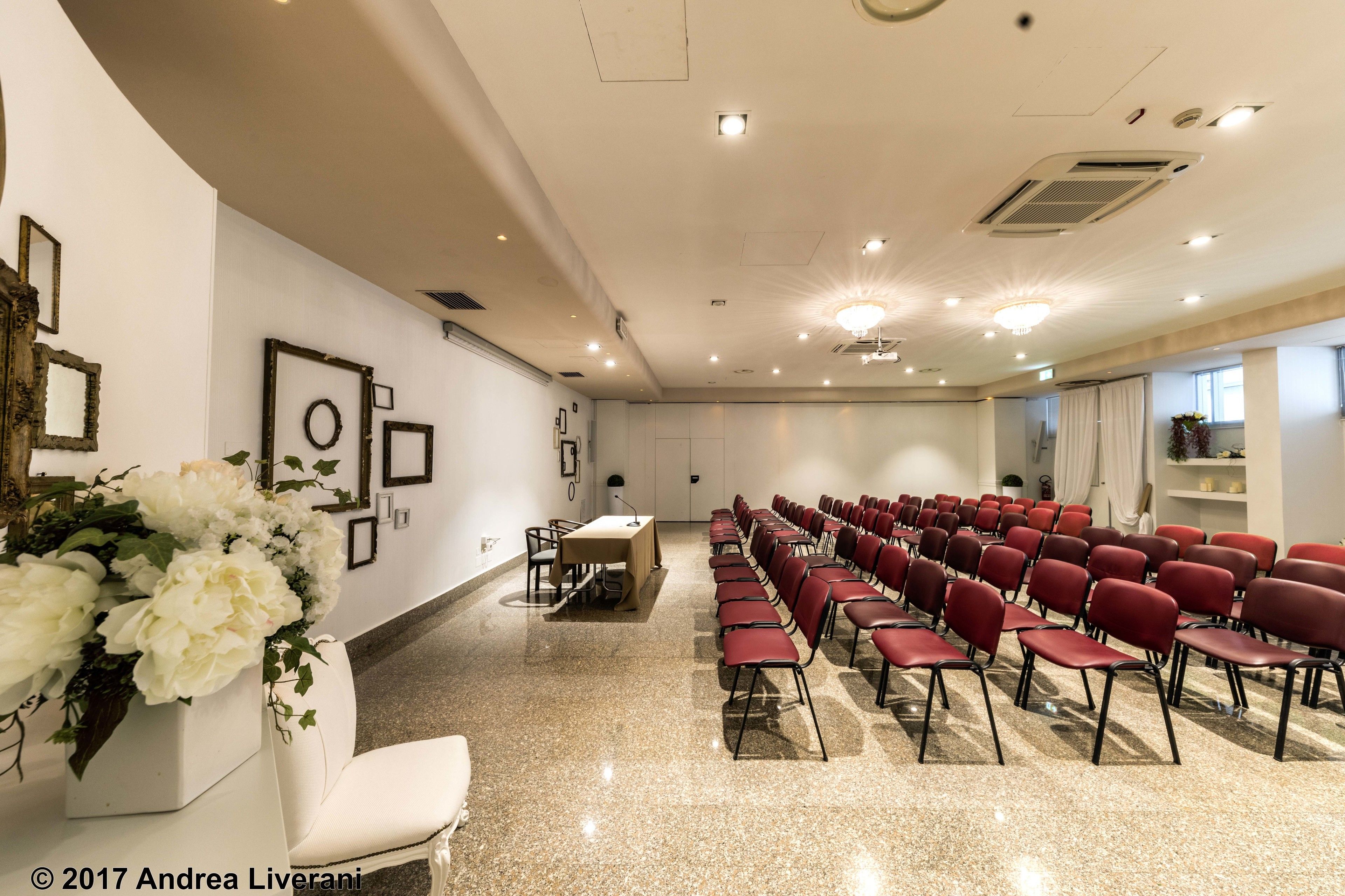 Une salle de conférence avec des rangées de chaises rouges, une table blanche et des tableaux encadrés accrochés aux murs.