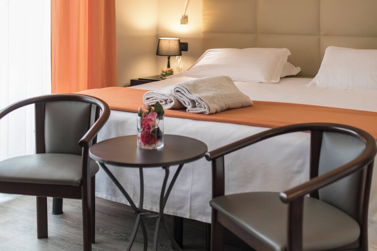 A hotel room with a bed, two chairs, a small table with a vase, and a nightstand with a lamp.