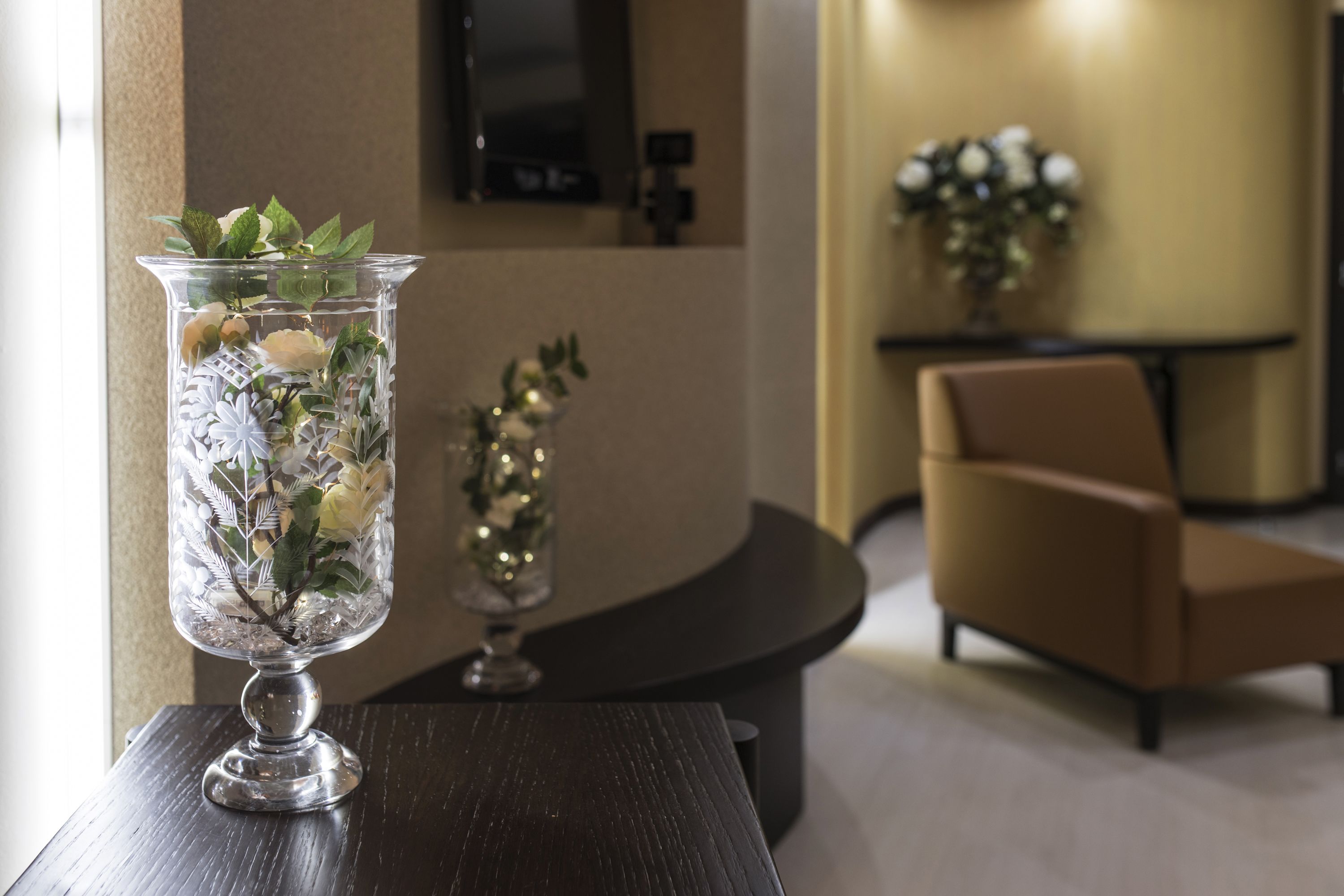 Un florero de vidrio transparente con flores sobre una mesa de madera oscura en una habitación moderna, con una silla de cuero marrón y una mesita con un objeto decorativo.