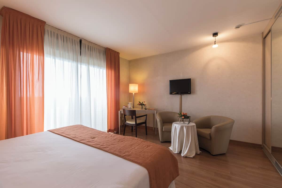 A hotel room with a bed, a desk with a chair, a small table with a chair, a TV mounted on the wall, and a window with curtains.