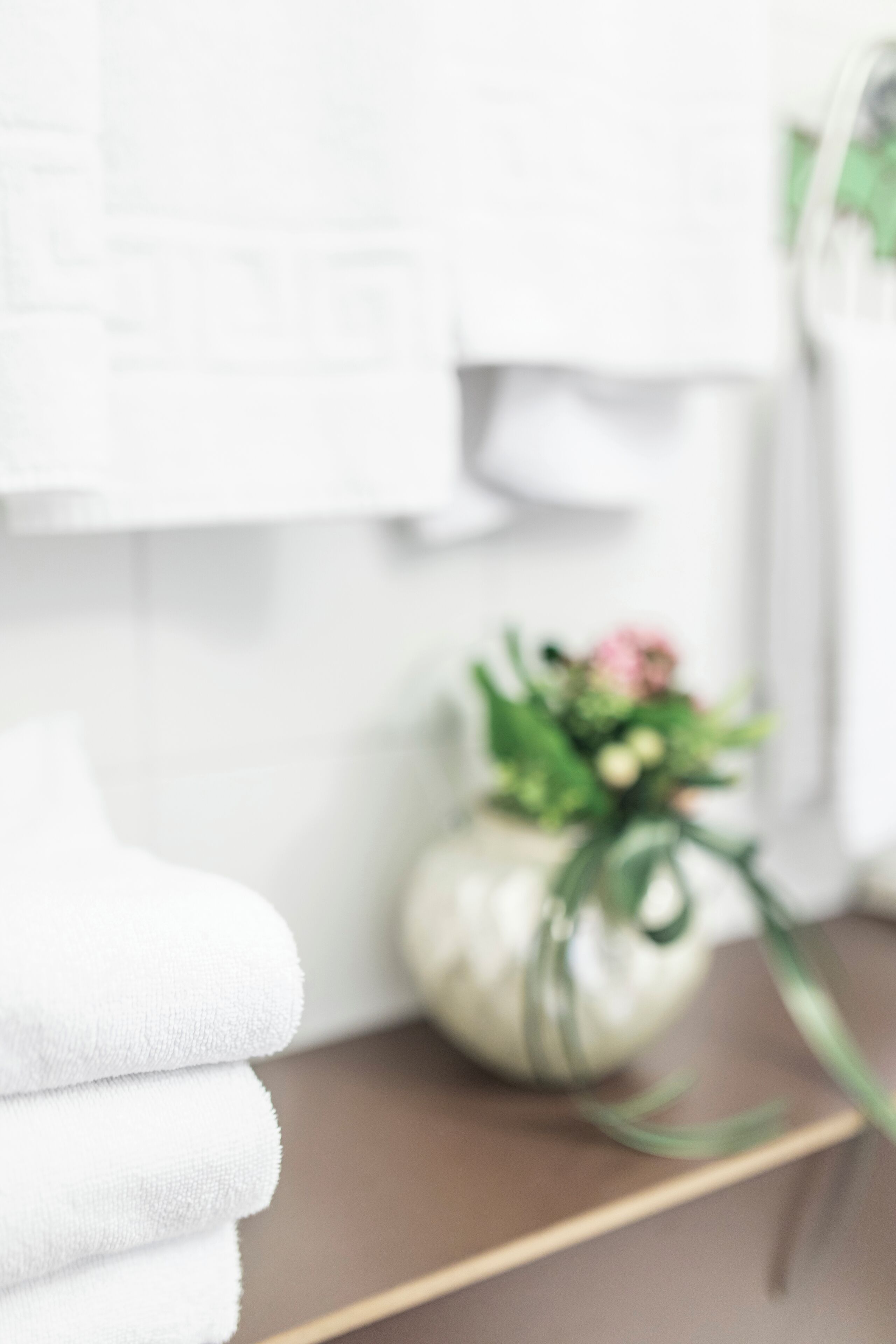 A stack of white towels on a wooden surface, a vase with flowers, and a white wall in the background.