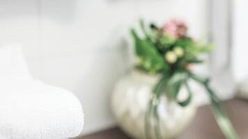 A stack of white towels on a wooden surface, a vase with flowers, and a white wall in the background.