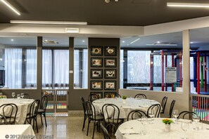 Un interior de restaurante con mesas redondas, sillas, cuadros en la pared y vistas a un parque infantil a través de las ventanas.