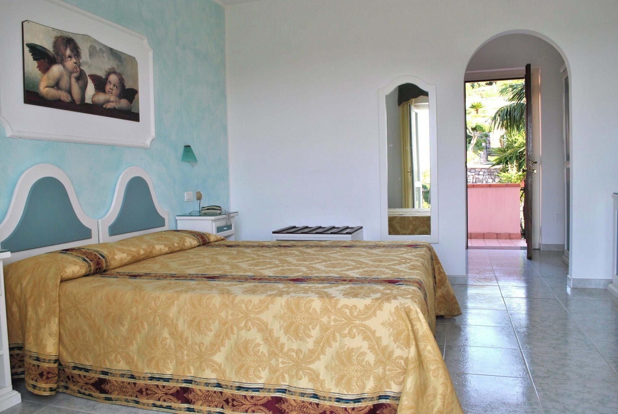 A hotel room with two beds, a painting on the wall, a bedside table with a lamp, and a view of the outside.