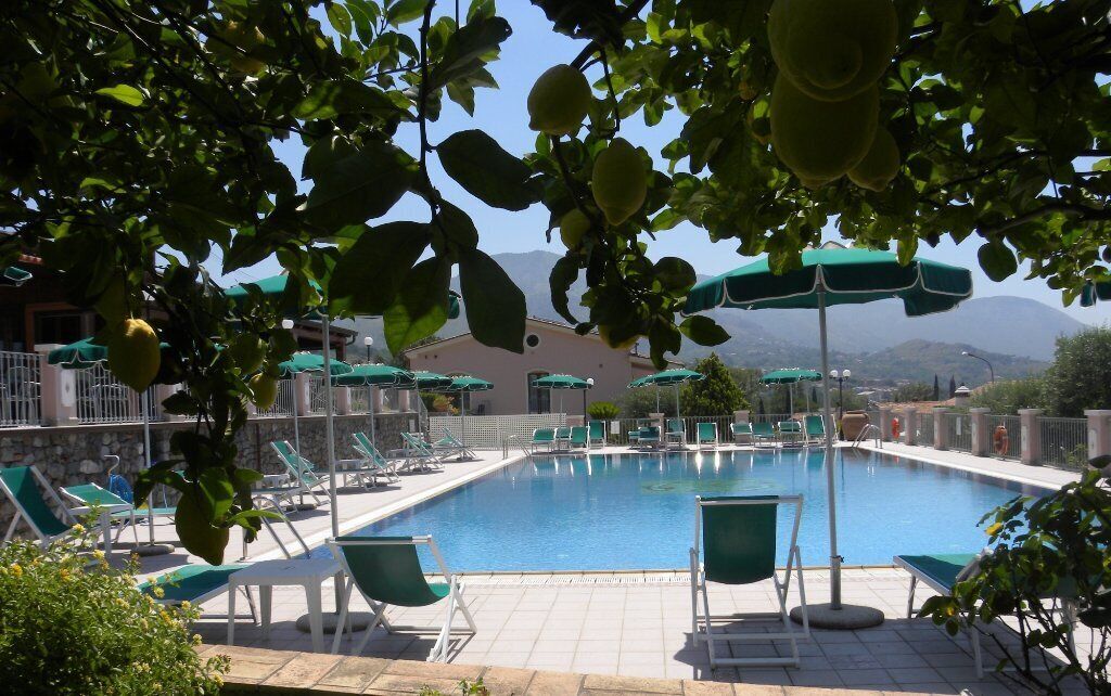 A swimming pool with green chairs and umbrellas, surrounded by a stone wall and trees.