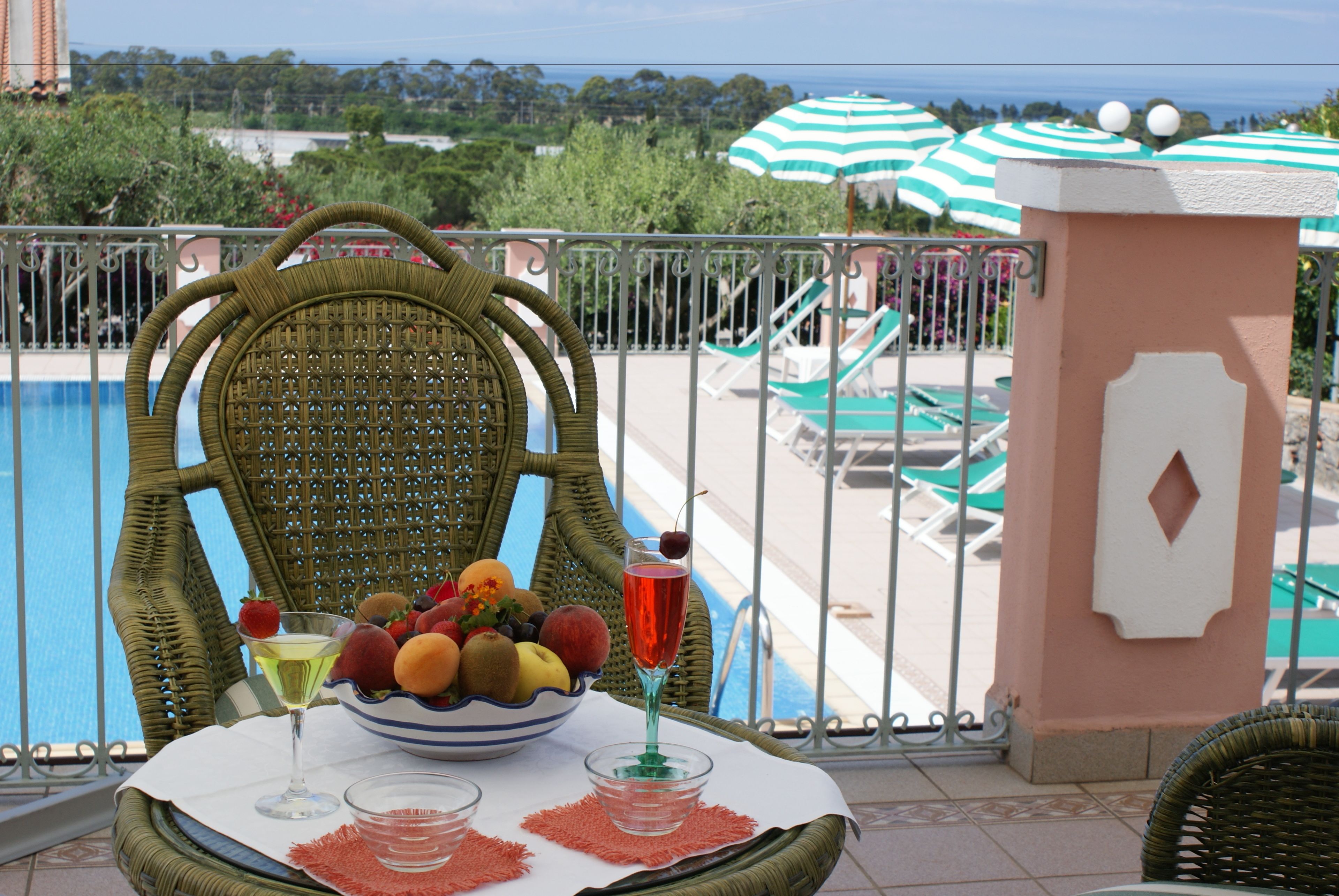 Una silla de mimbre con un cuenco de frutas y bebidas en un balcón con vistas a una piscina y sillas de descanso.