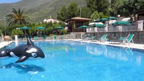 Piscina com uma grande orca inflável, rodeada por espreguiçadeiras e guarda-sóis, com montanhas ao fundo.