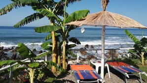 Um espaço de descanso à beira-mar, com espreguiçadeiras e um guarda-sol de palha, com vista para o oceano.