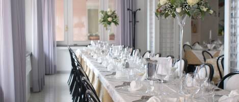 A long dining table set with white tablecloths, silverware, and clear glassware, flanked by black chairs.