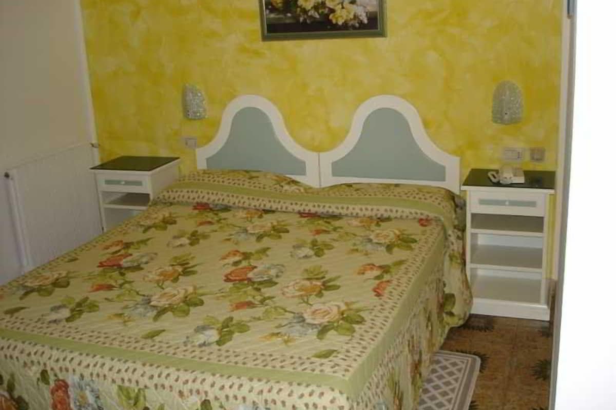 A bedroom with a floral bedspread, a white headboard, a nightstand with a lamp, and a bedside table.