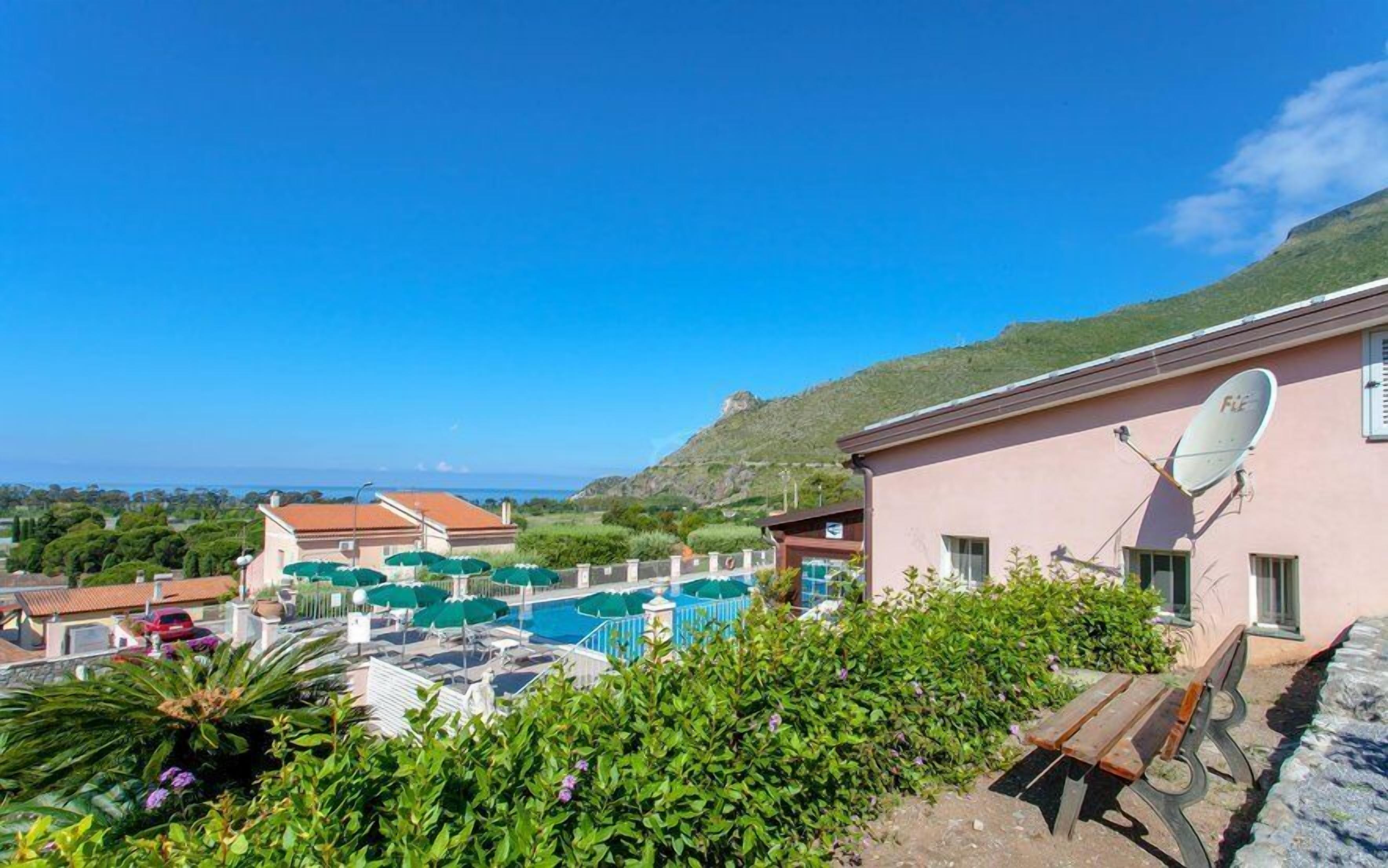 A pool area with a view of the sea, surrounded by greenery and a building with a satellite dish.