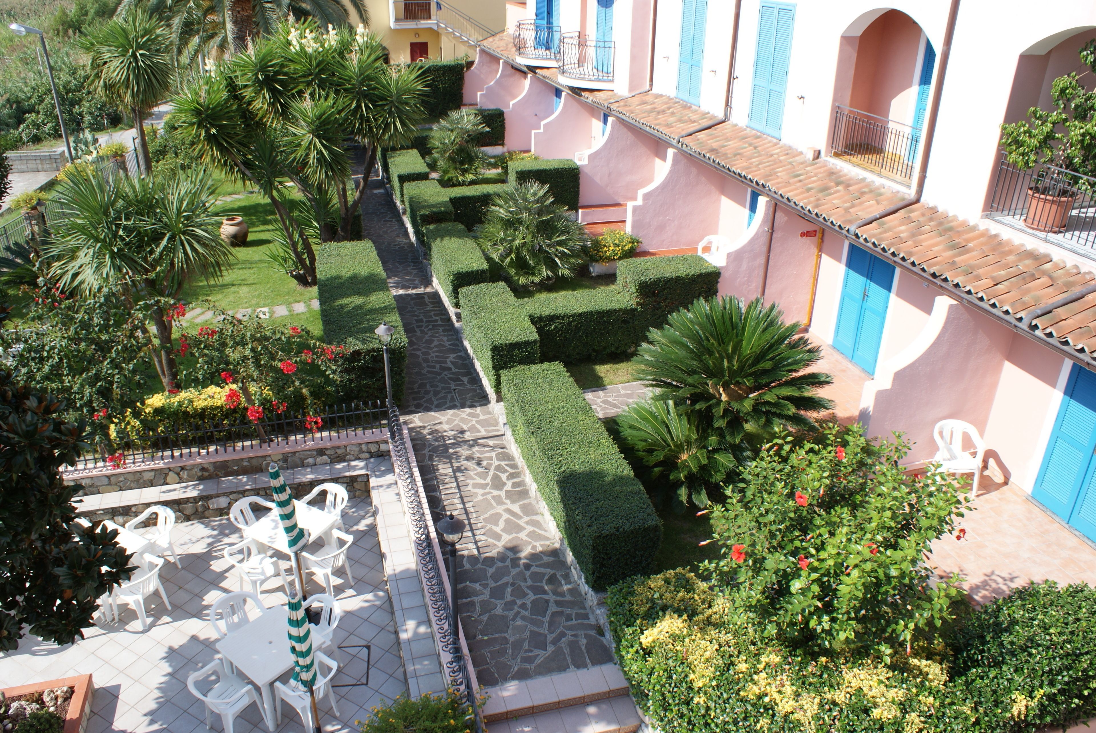 A courtyard with neatly trimmed hedges, a stone pathway, and outdoor seating with white chairs and a striped umbrella.