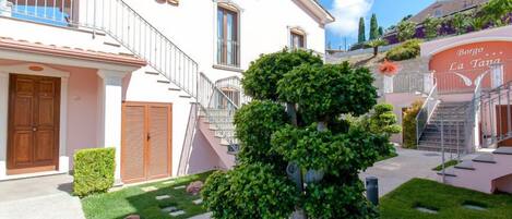 Un edificio de dos pisos con una escalera que conduce a un balcón, un pequeño jardín con un árbol cuidado, y un letrero que dice “Borgo La Tana”.