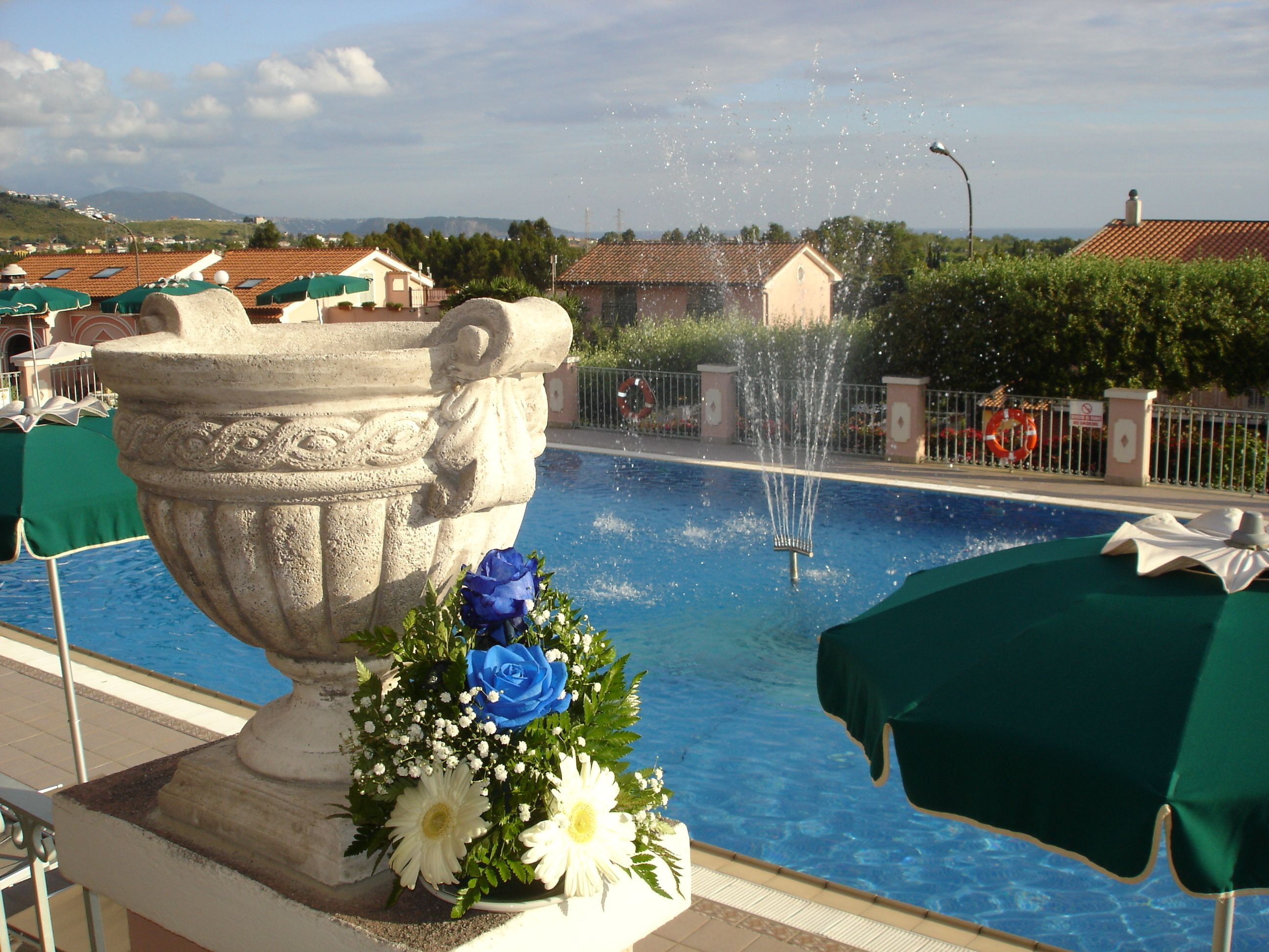 A pool with a fountain, surrounded by a stone urn with flowers, and a view of houses in the background.