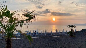 Uma praia com palmeiras, uma fileira de cadeiras de praia e guarda-sóis sob um céu ao pôr do sol.