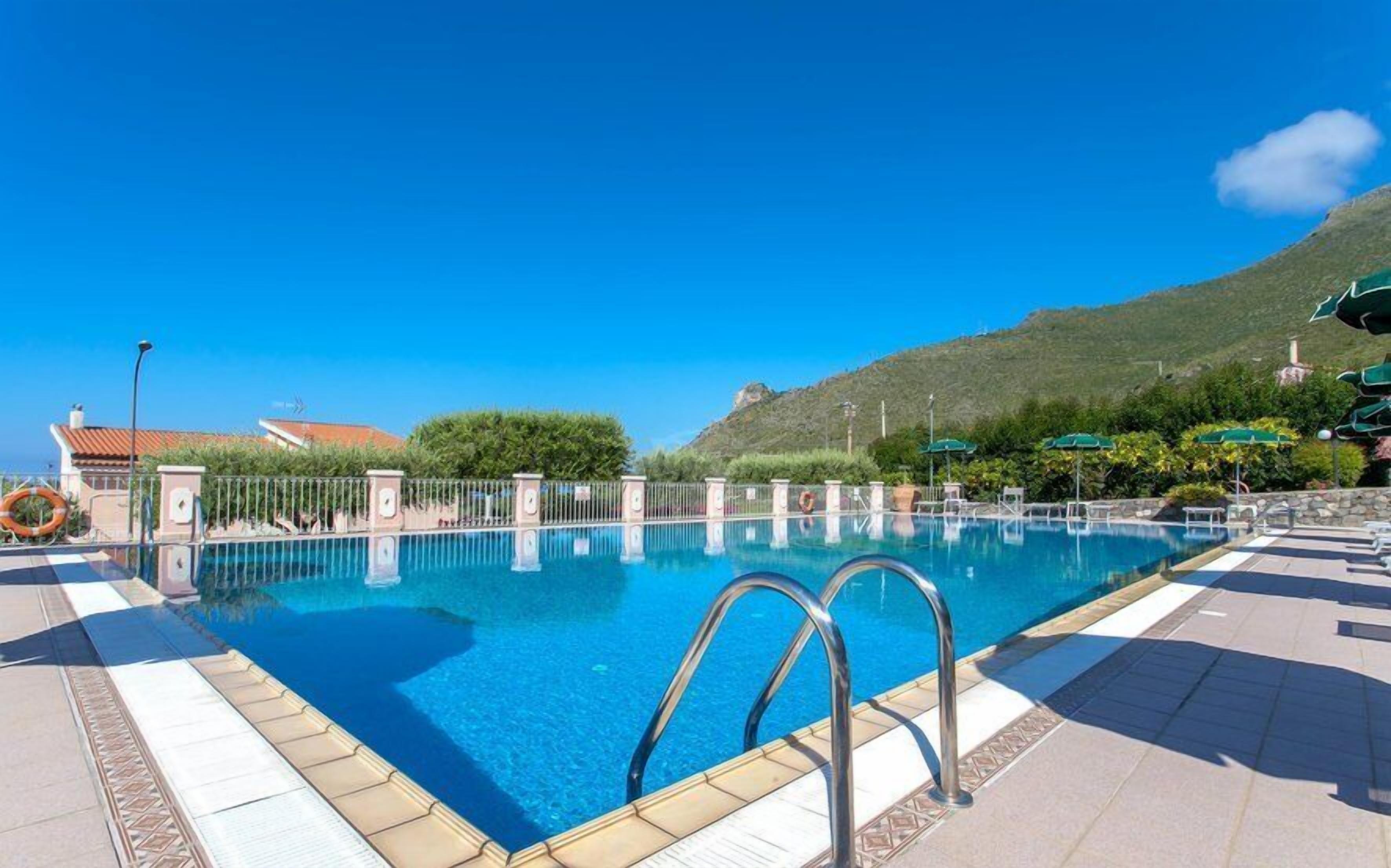 A clear blue pool with a metal ladder, surrounded by a tiled deck and a fence.