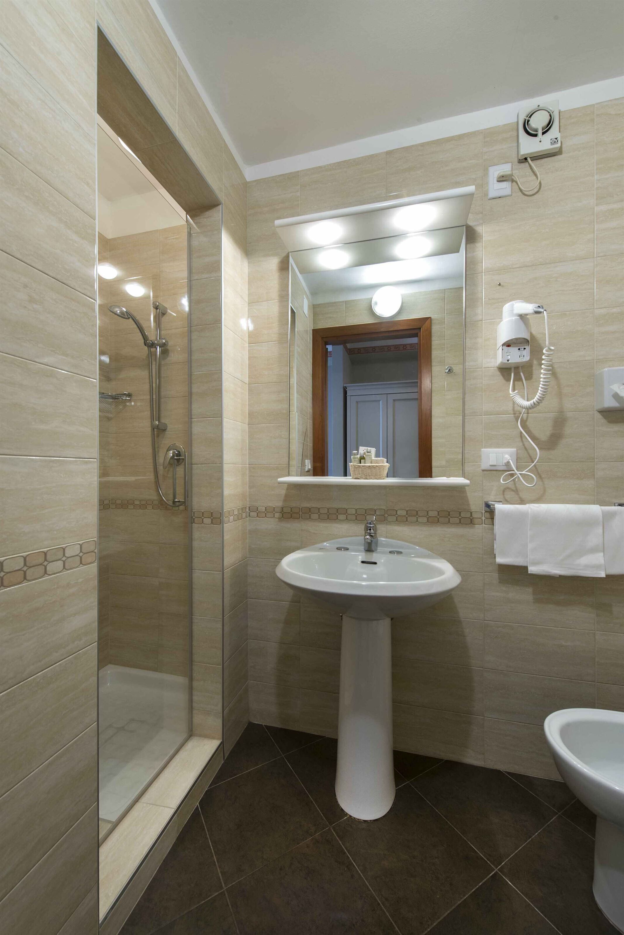A bathroom with a glass-enclosed shower, a pedestal sink, a mirror, and a hairdryer mounted on the wall.