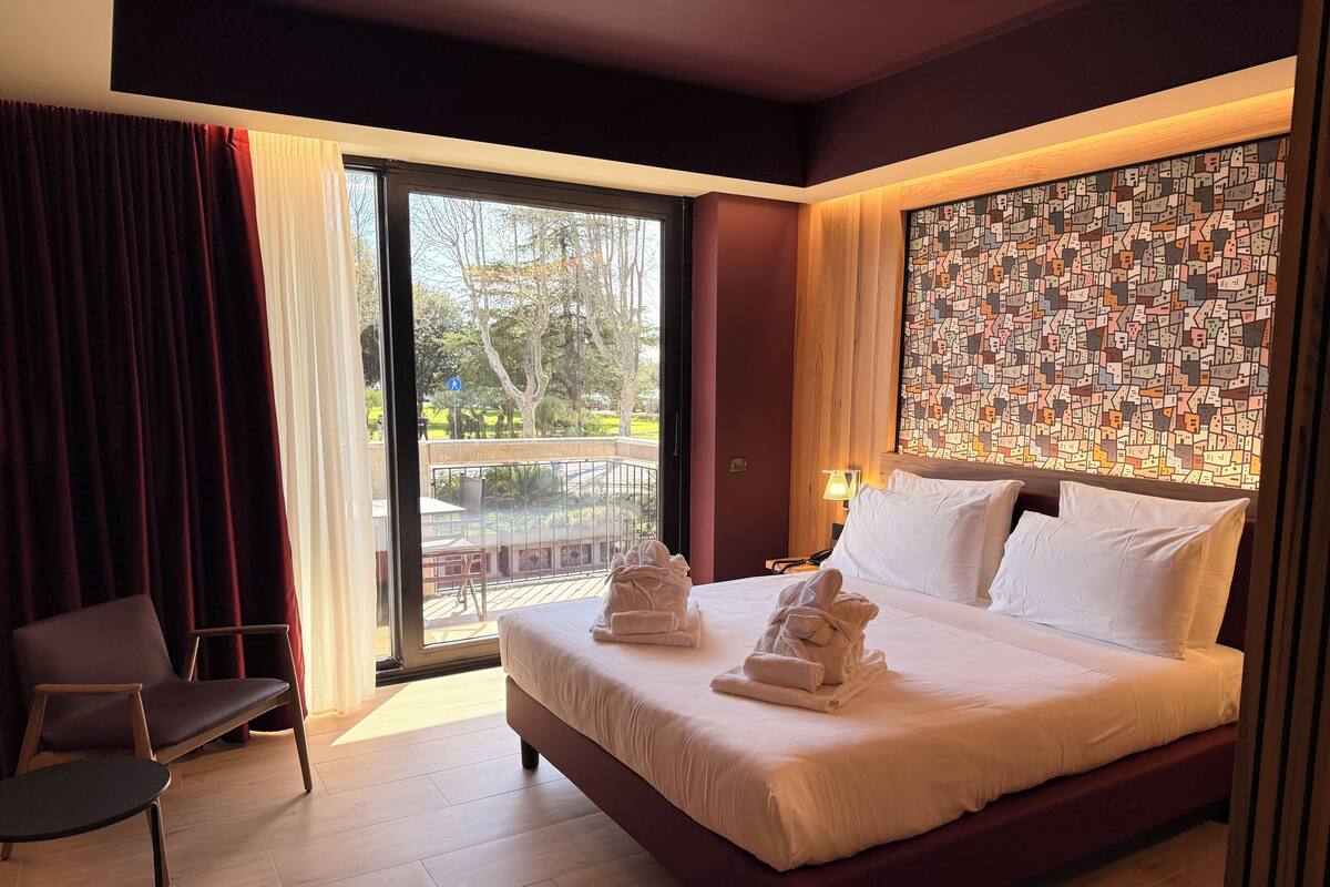 Ein Hotelzimmer mit einem großen Bett, einem Sessel, Blick ins Freie und einer Wand mit einer Weltkarten-Musterung.
