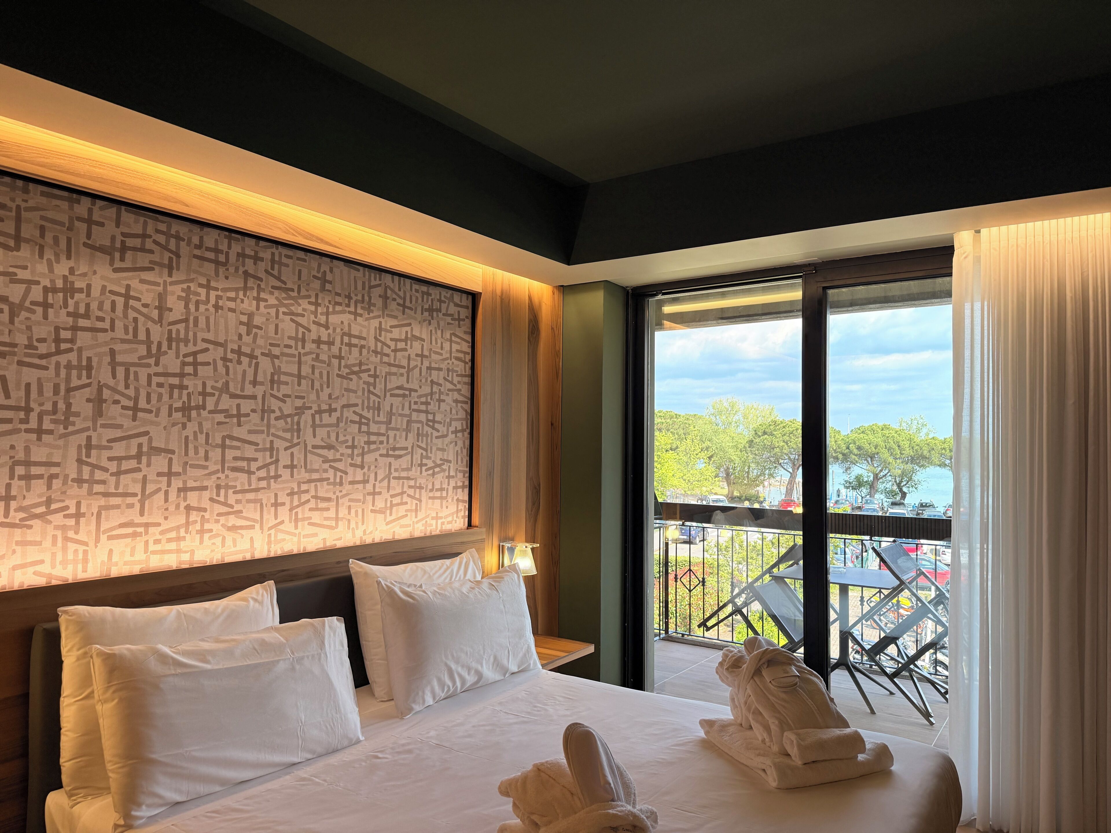 A hotel room with a bed, pillows, a headboard, a window with curtains, and a view of the outside.
