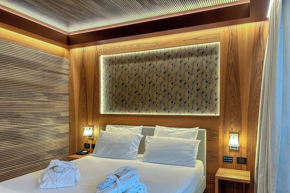 Ein Hotelzimmer mit einem ordentlich bezogenen Bett, einem Nachttisch mit Lampe und einer Wand mit geometrischem Muster.