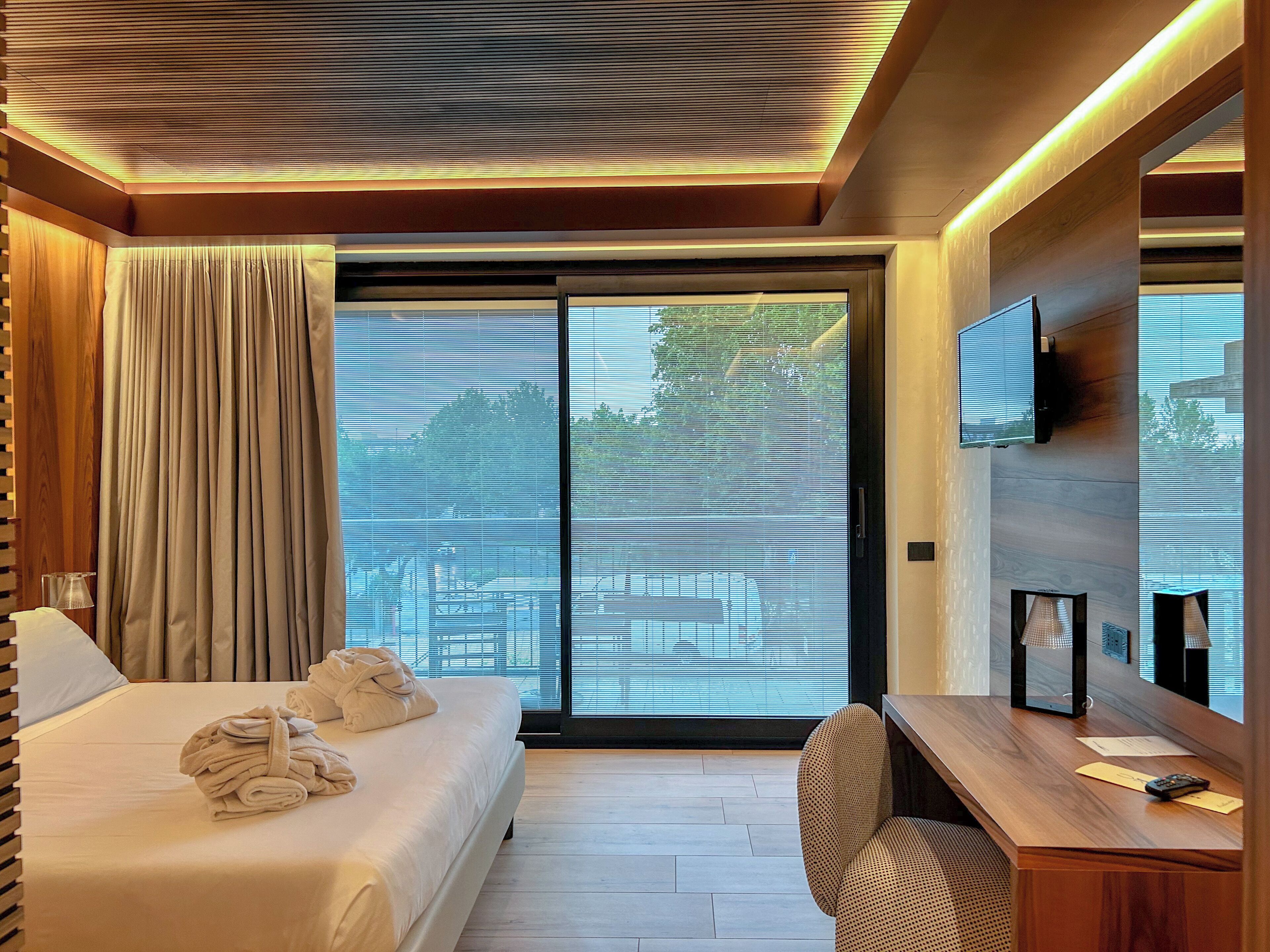 Habitación de hotel con cama, escritorio, silla, televisor montado en la pared y un amplio ventanal corredizo.