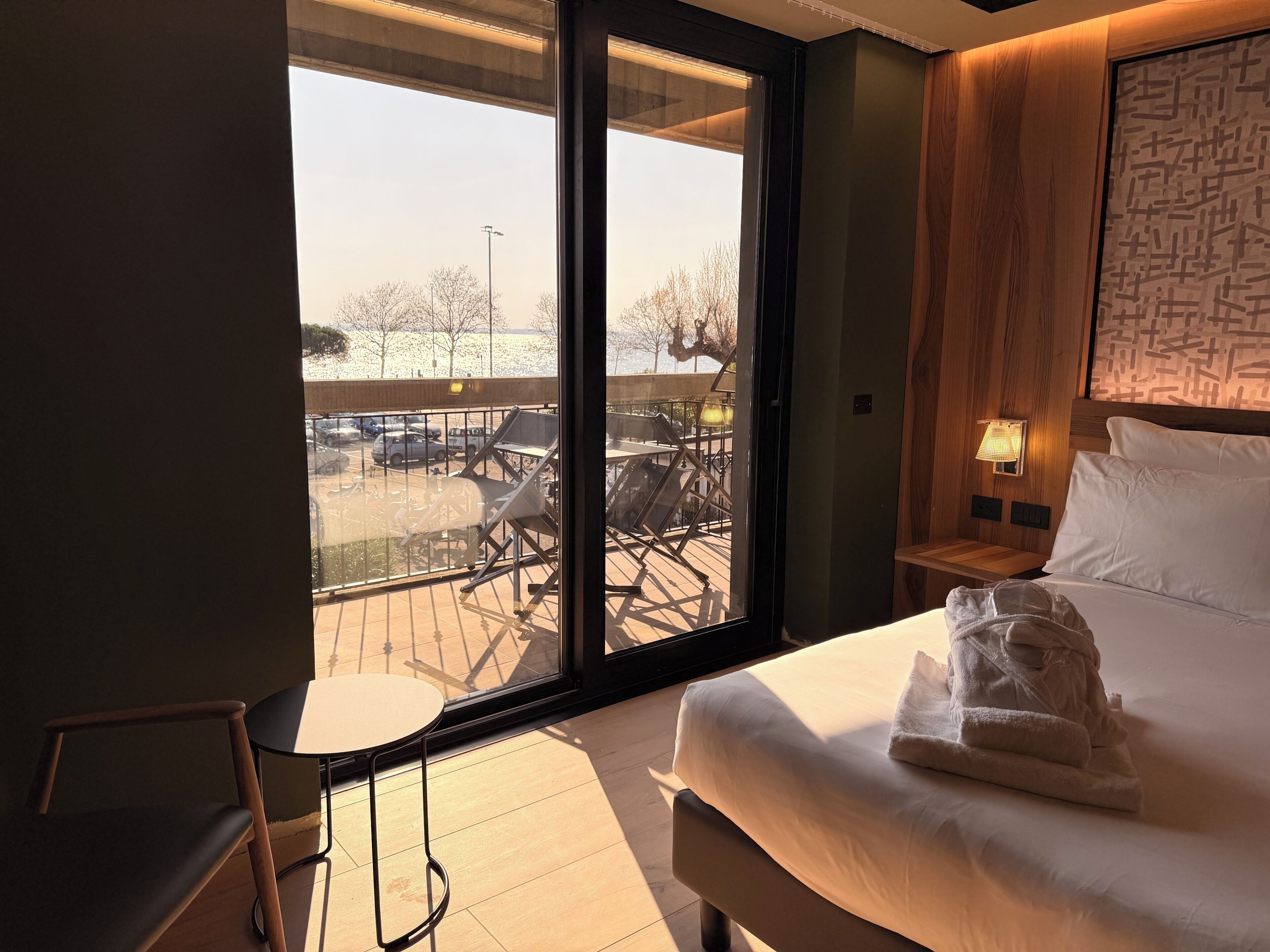 A hotel room with a large sliding glass door to an outdoor area, a bed with white linens, a small bedside table with a lamp, and a wooden headboard.