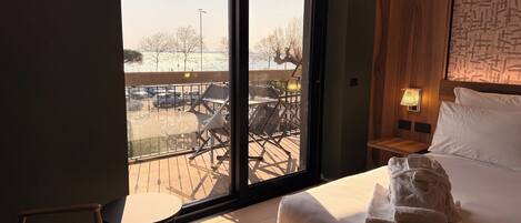 A hotel room with a large sliding glass door to an outdoor area, a bed with white linens, a small bedside table with a lamp, and a wooden headboard.