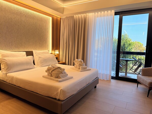 A modern hotel room with a large bed, a nightstand with a lamp, a balcony with a chair, and a view of greenery outside.