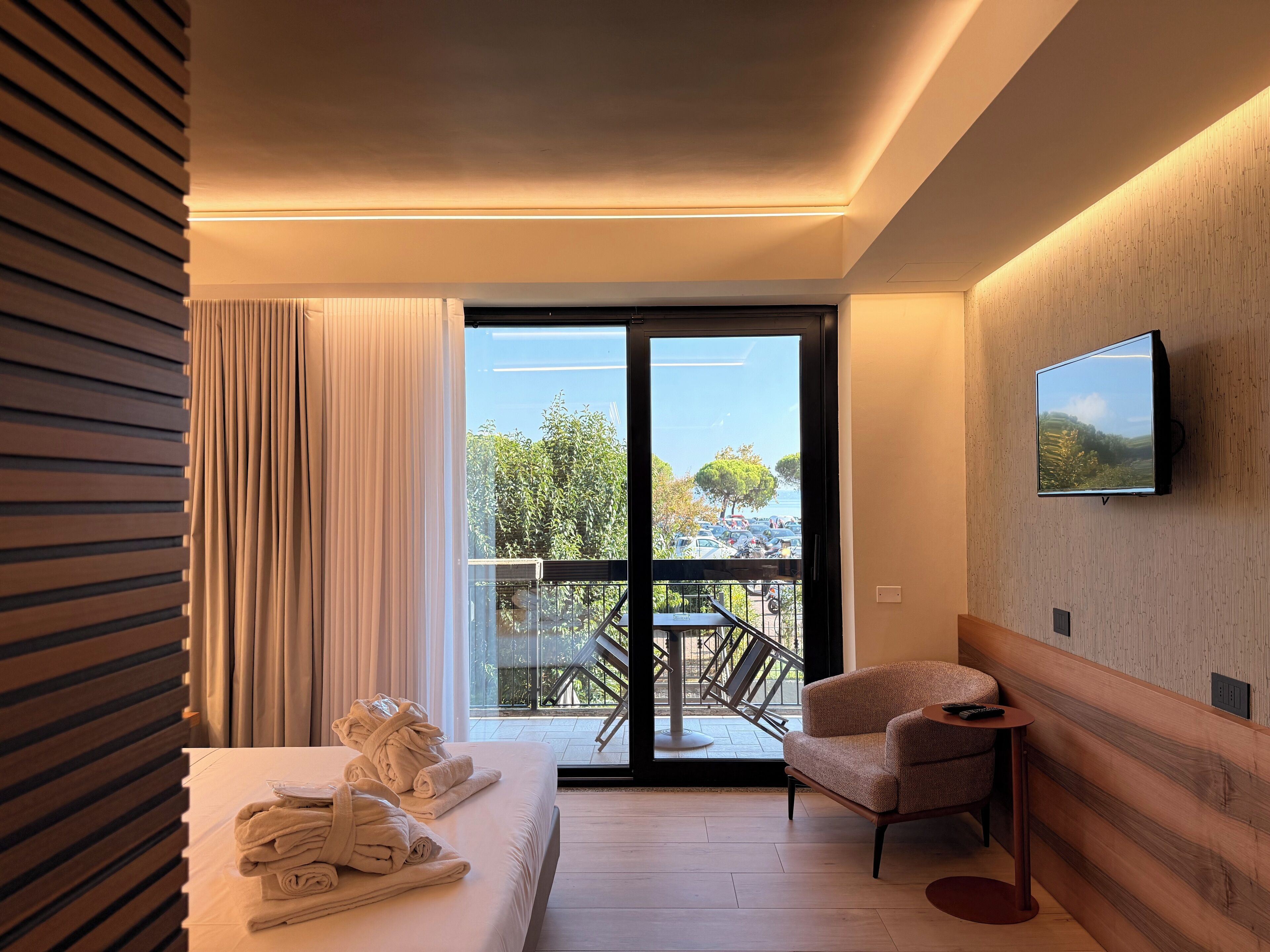 Una moderna camera d'albergo con un letto, una sedia, una TV e un balcone con vista su spazi verdi e auto parcheggiate.