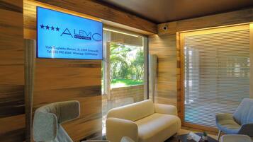 Una habitación de hotel moderna con una televisión grande mostrando información del hotel, un sofá, dos sillones y una mesita con una planta.