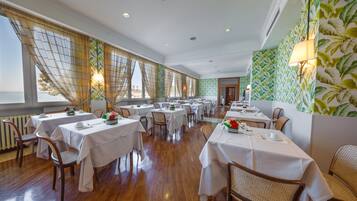 Un ristorante con diversi tavoli e sedie, ampie finestre con tende e una parete decorata con un motivo a foglia.