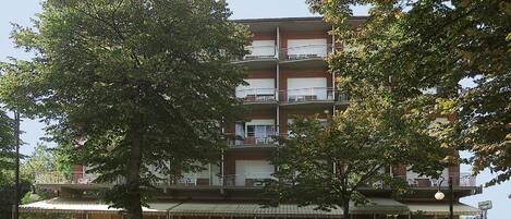 Un edificio de varios pisos con balcones, un área de estar al aire libre techada y un árbol en primer plano.