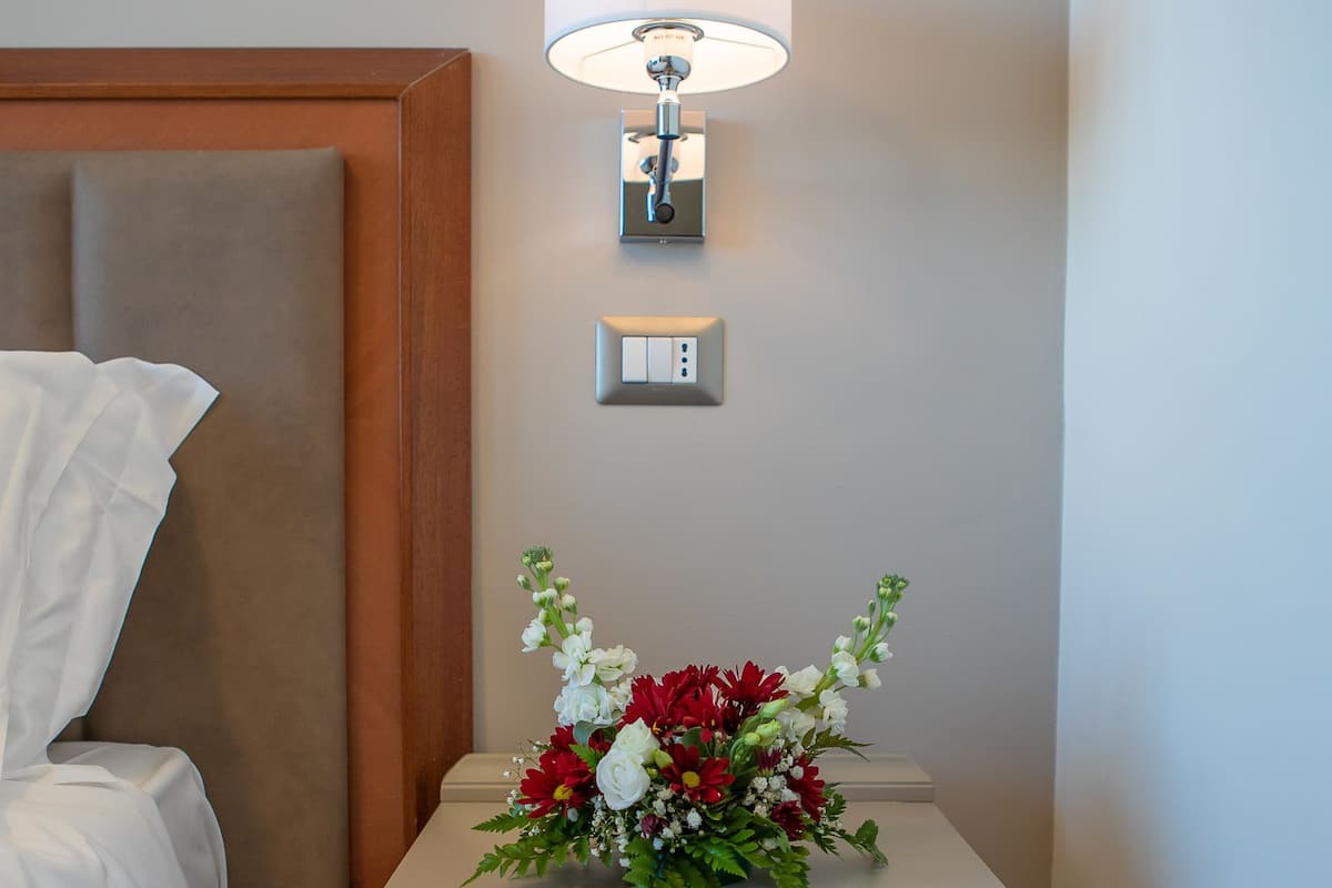 En snyggt bäddad säng med ett vitt täcke och kuddar, ett sängbord med en lampa och en blomsteruppsättning samt en sänggavel i trä.