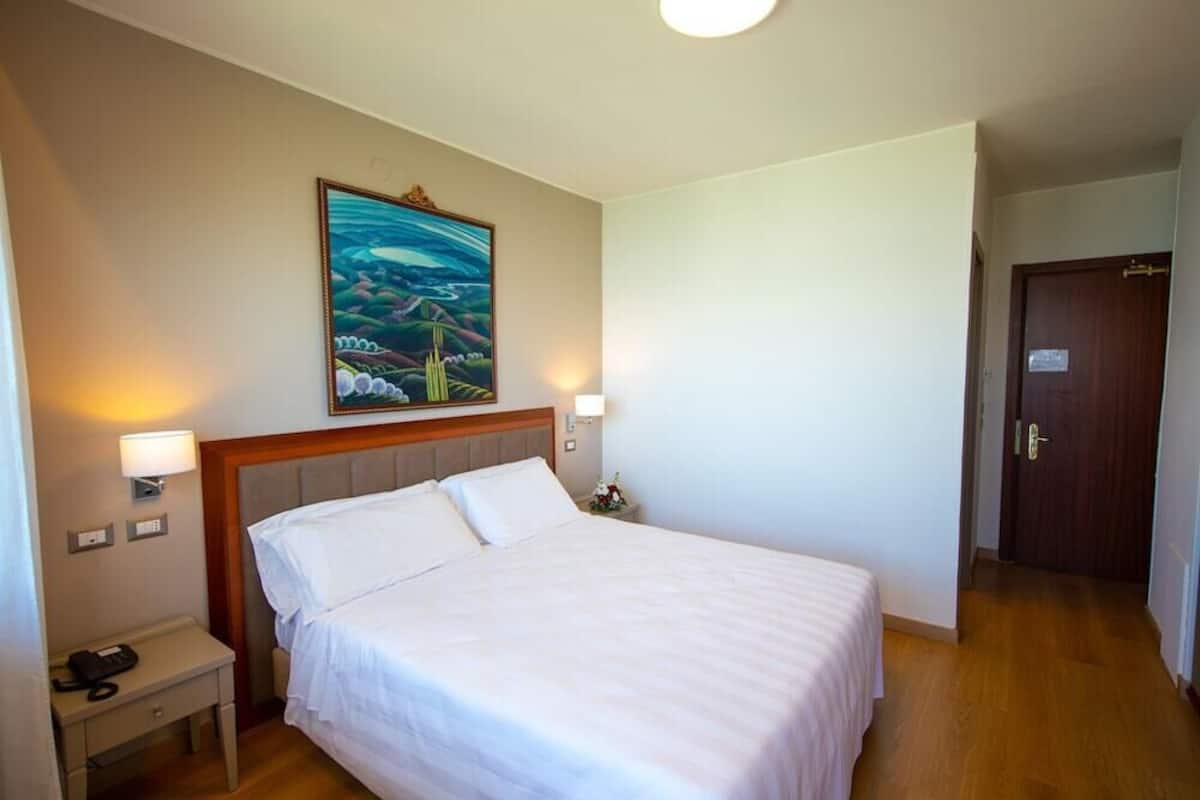Une chambre d’hôtel avec un lit, des lampes de chevet, une table de nuit, un téléphone, un tableau au mur et une porte.