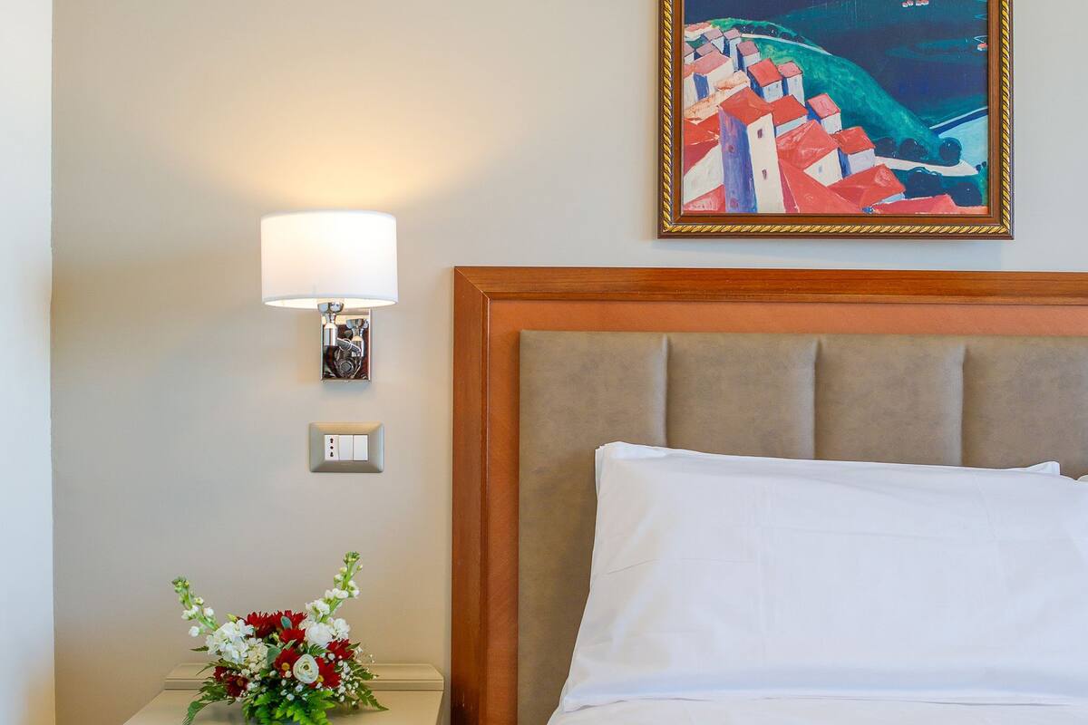 Ett hotellrum med en säng, ett nattduksbord, en lampa, en tavla och en blomsteruppsättning.