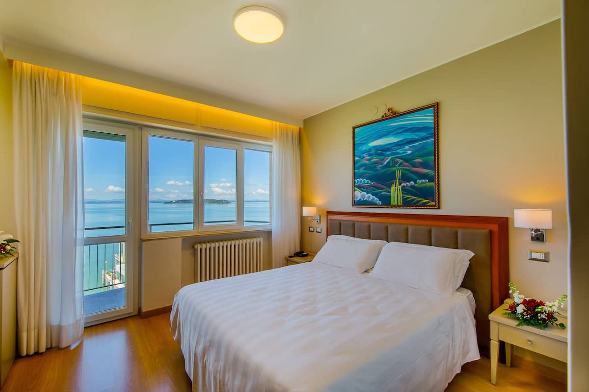 Ett hotellrum med en stor säng, utsikt över havet genom ett fönster och en tavla på väggen.