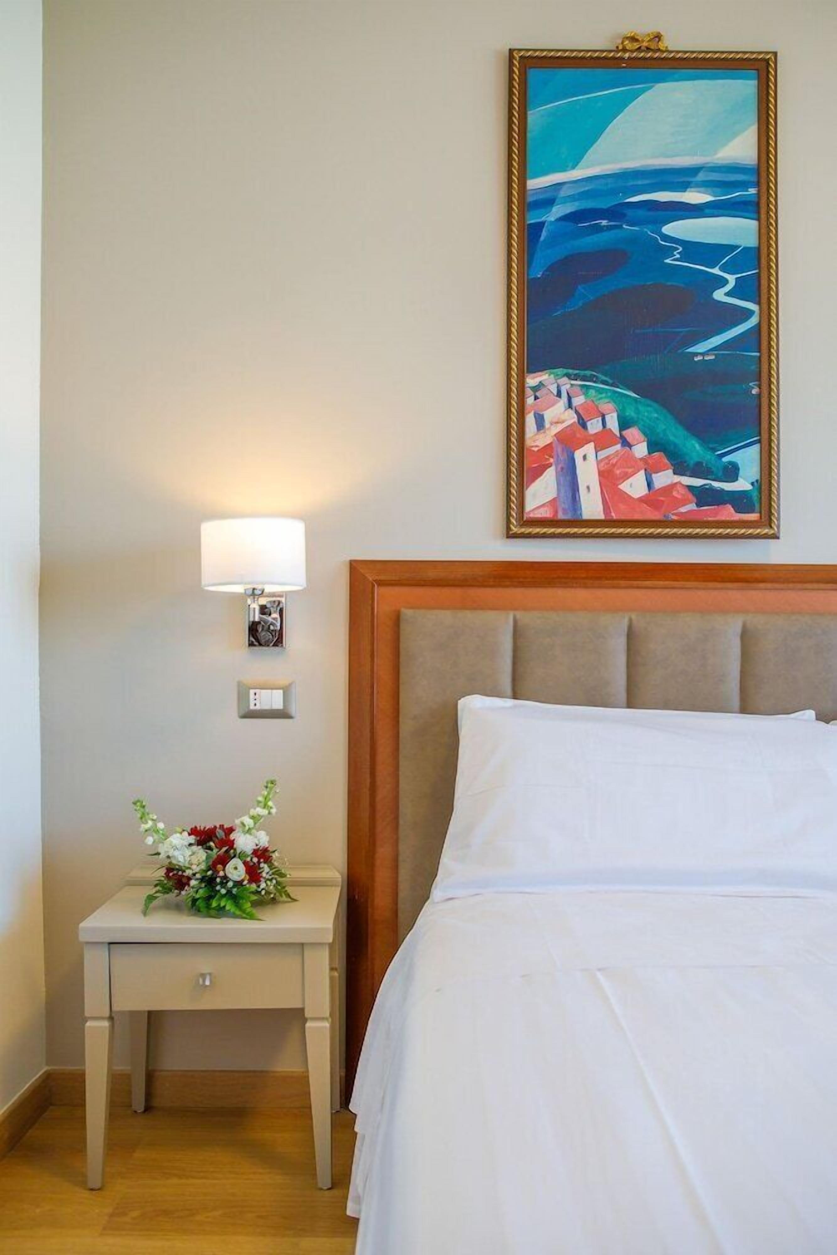 Ein Hotelzimmer mit Bett, Nachttisch mit Lampe, einem gerahmten Gemälde und einer Blumenkomposition.