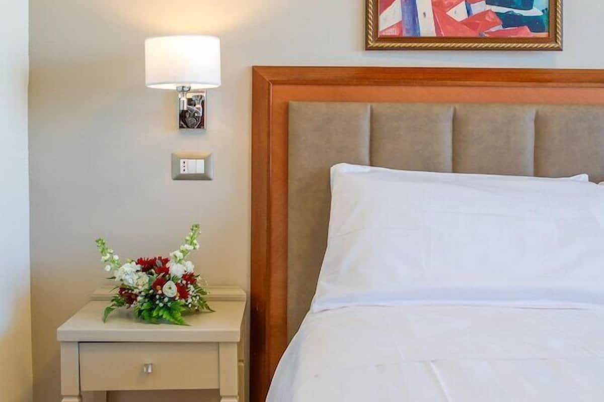 Ett hotellrum med en säng, ett nattduksbord med en lampa, en inramad tavla och en blomsteruppsättning.